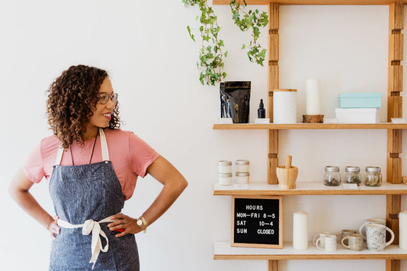 Woman in a pink shirt and blue apron standing in a store with shelves displaying various items.